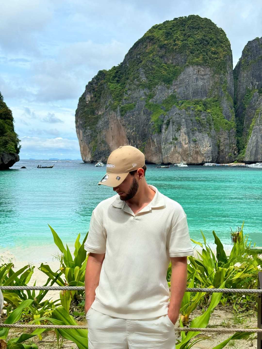 O Vagner de boné e roupa branca, de pé, na frente de uma baía de água turquesa entre paredões de pedra.