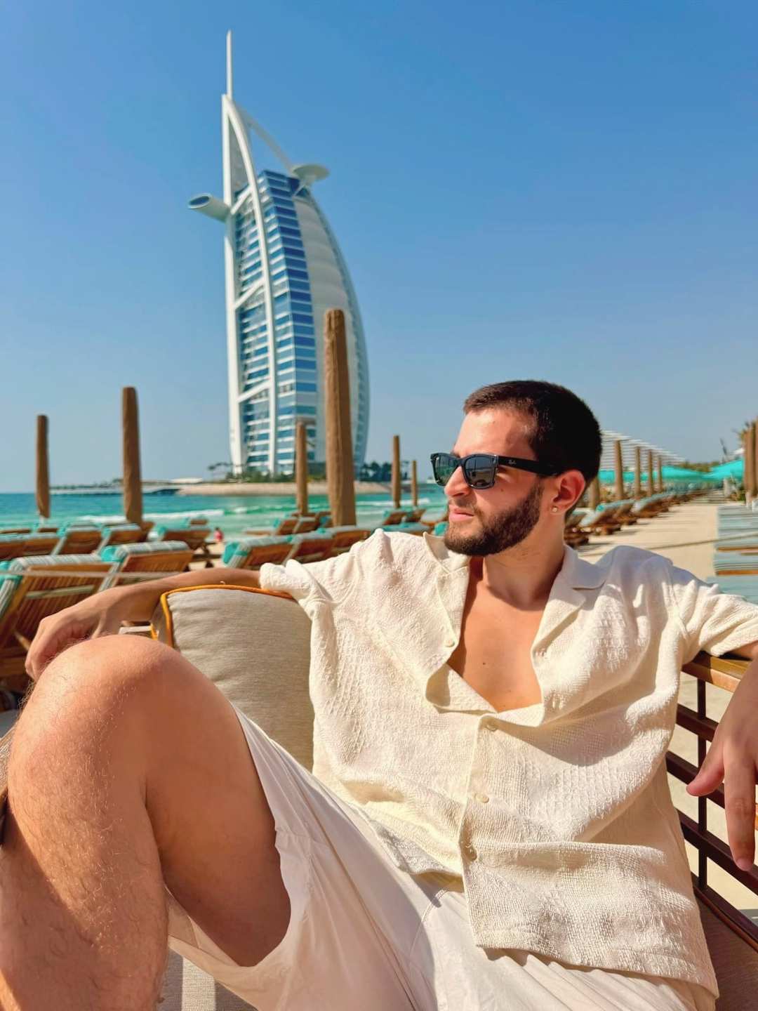 O Vagner sentado à beira-mar em Dubai, de óculos escuros, com o hotel em forma de vela atrás.