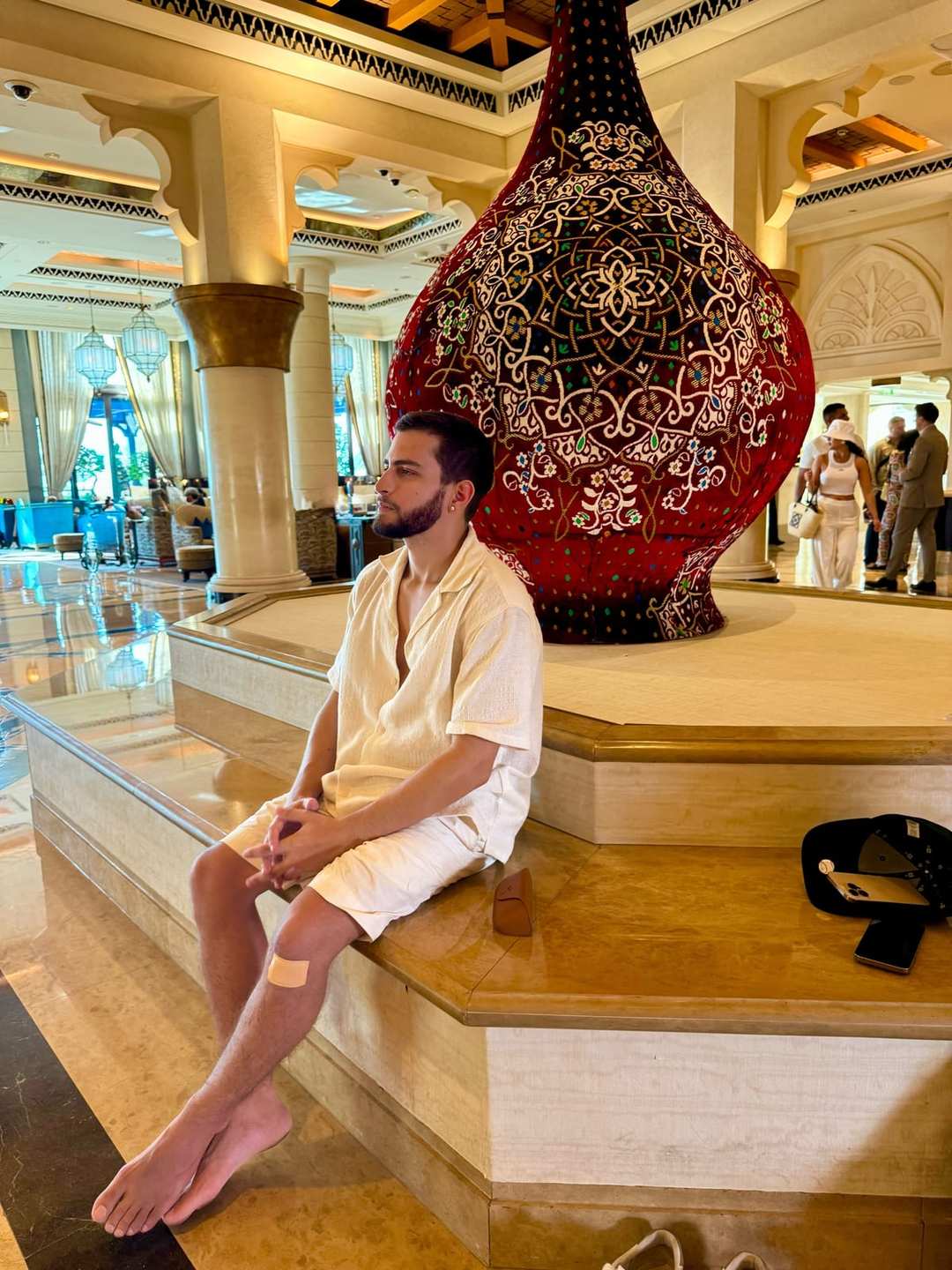 O Vagner sentado no saguão de um hotel em Dubai, de roupa branca, com um vaso gigante ornamentado atrás.