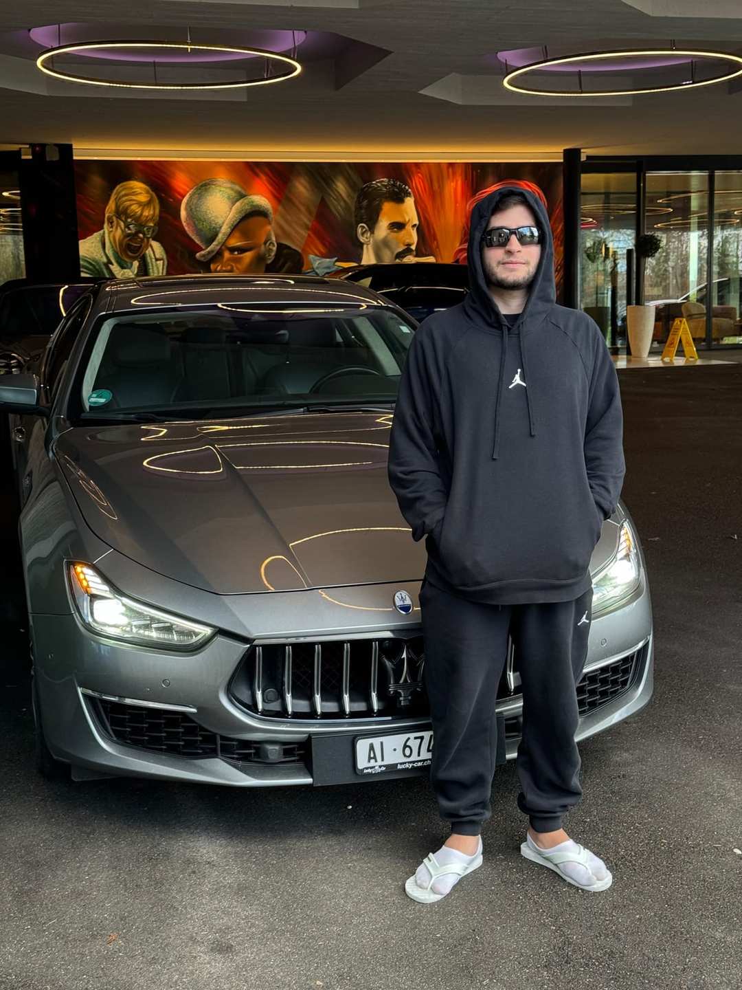 O Vagner de moletom preto, capuz e óculos escuros, de pé ao lado de uma Maserati cinza na entrada de um hotel na Suíça.