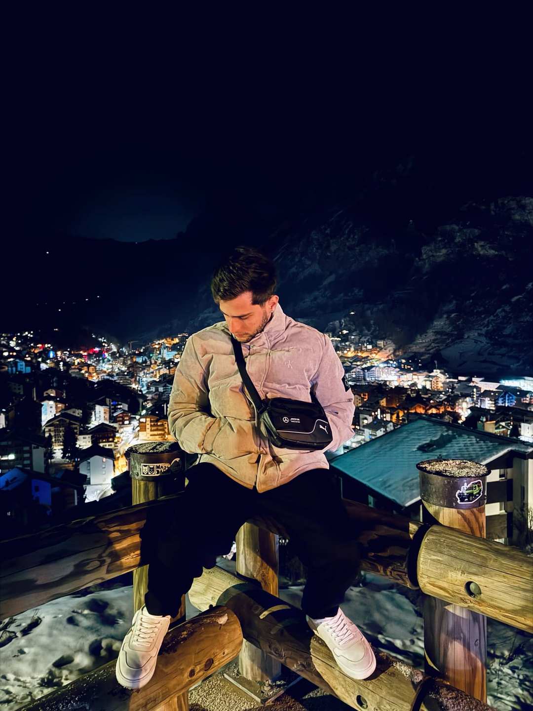 O Vagner sentado num mirante à noite, de jaqueta, com as luzes de uma cidade na encosta atrás.