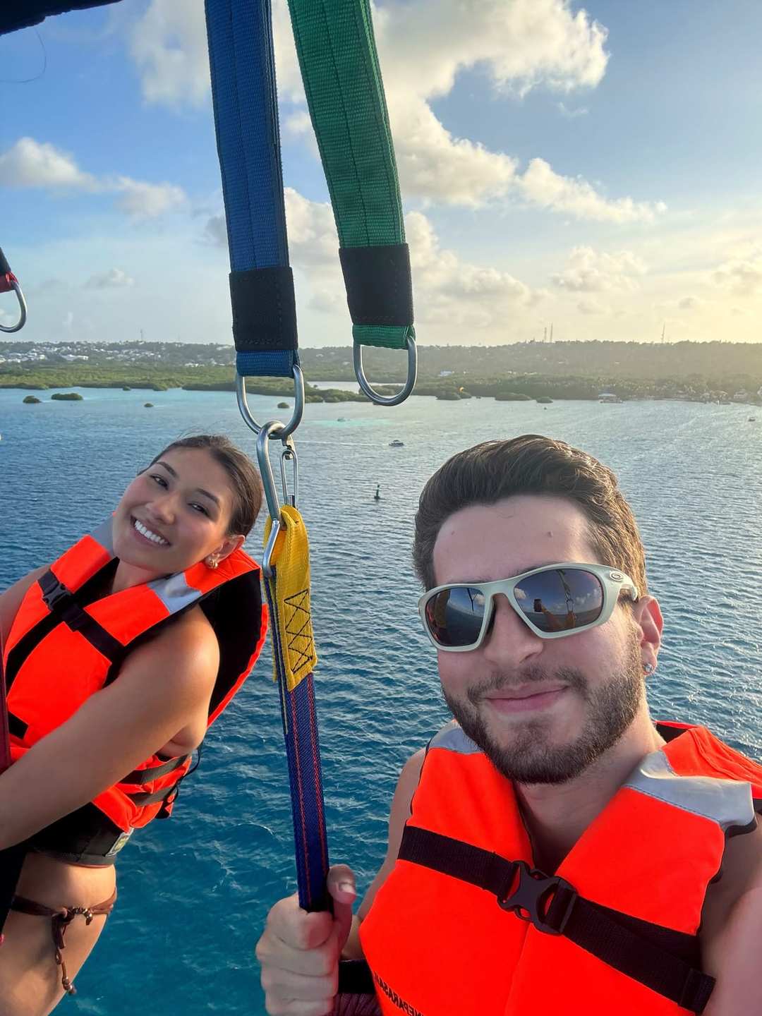 O Vagner e a mulher de colete salva-vidas laranja, pendurados num parasail sobre o mar.