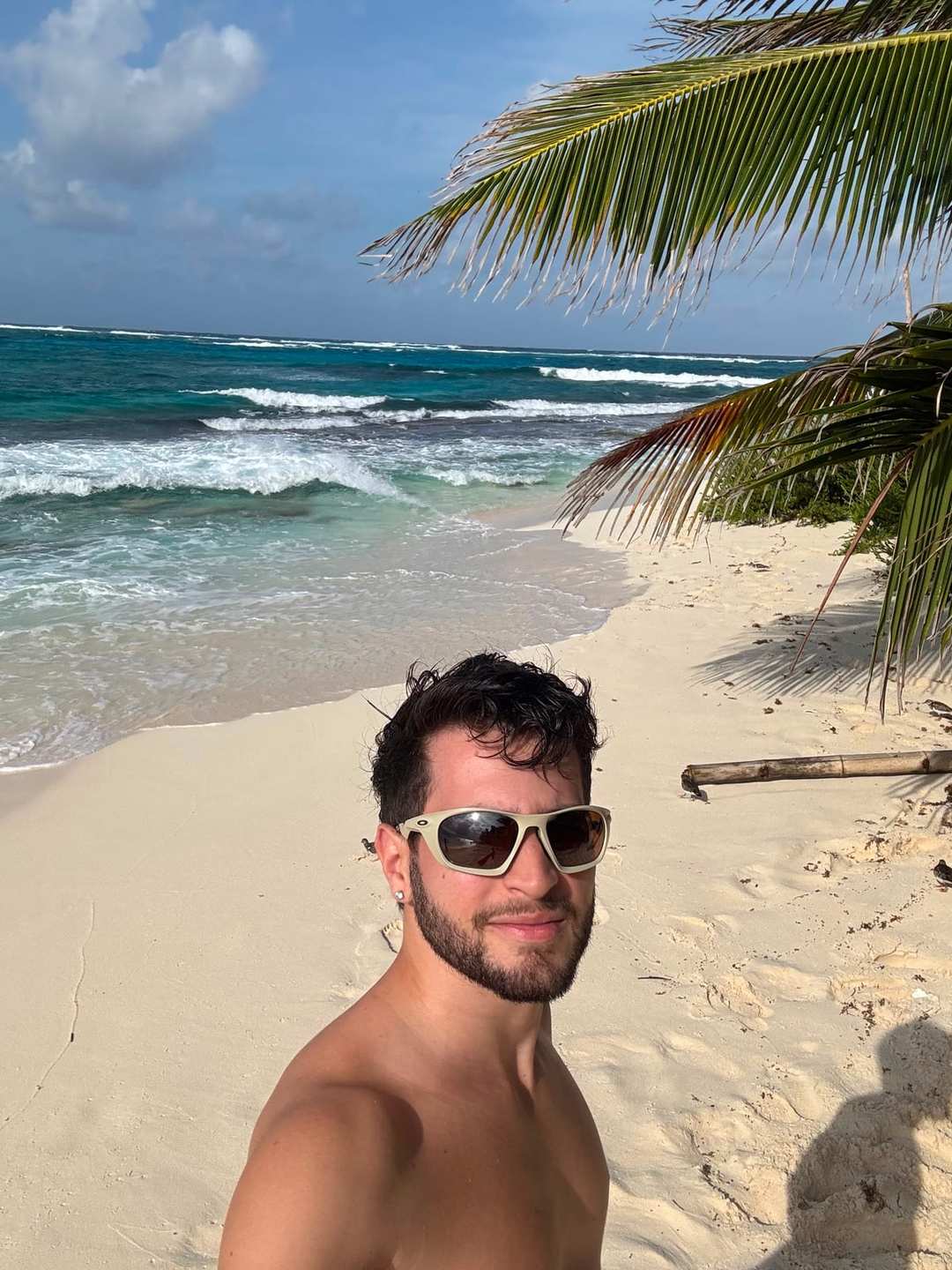 O Vagner sem camisa, de óculos escuros, numa praia de areia branca com coqueiros e o mar batendo atrás.