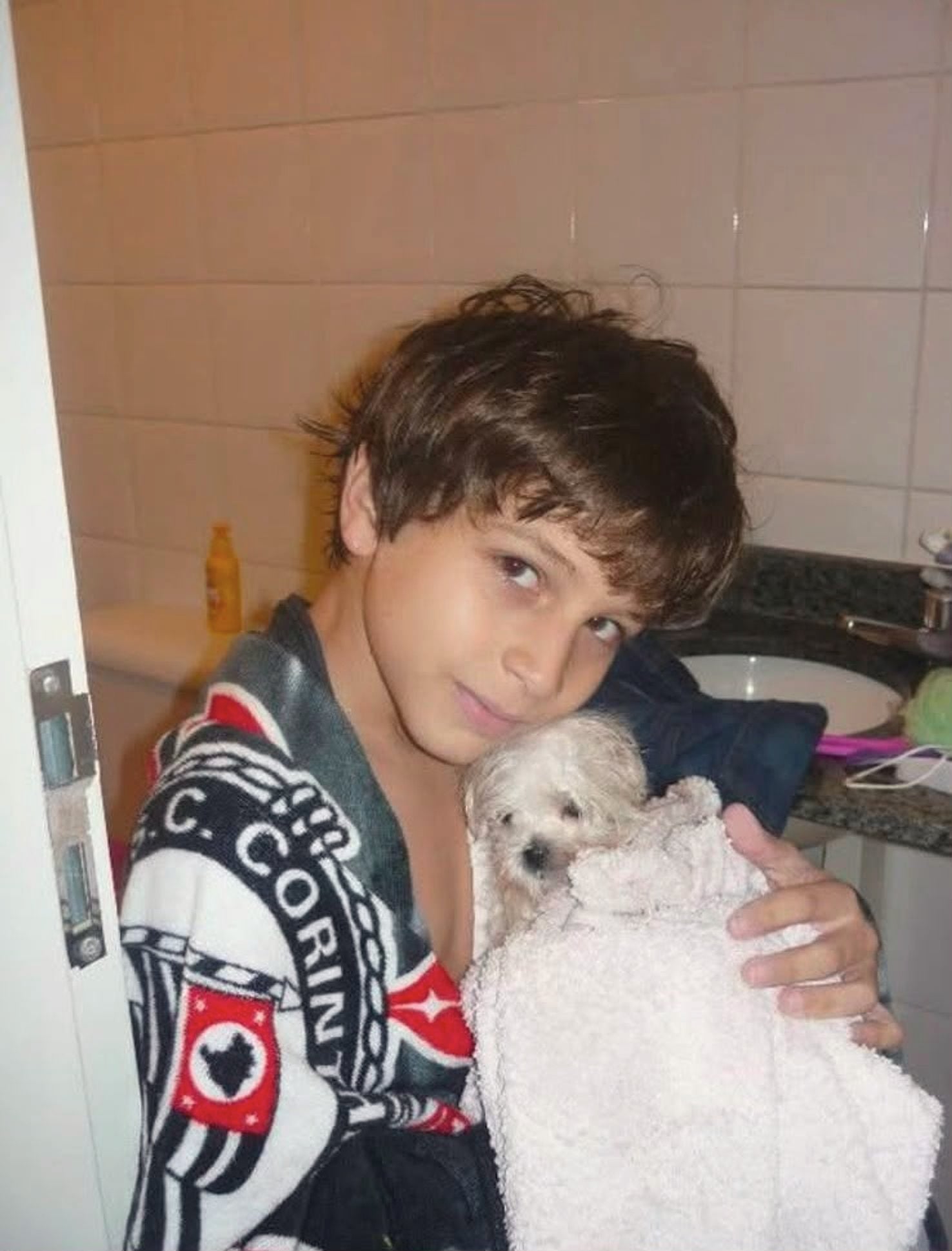O Vagner criança, num banheiro de azulejo, enrolado numa toalha do Corinthians, segurando um cachorrinho branco.