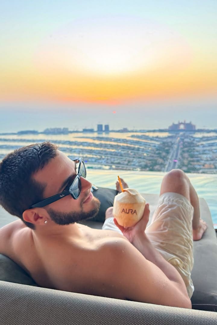 O Vagner deitado numa espreguiçadeira à beira de uma piscina no alto de um prédio em Dubai, com um coco na mão e o pôr do sol sobre a Palm Jumeirah.