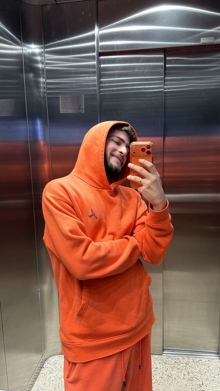 O Vagner de moletom laranja com o capuz na cabeça, sorrindo, tirando uma selfie no espelho de um elevador.