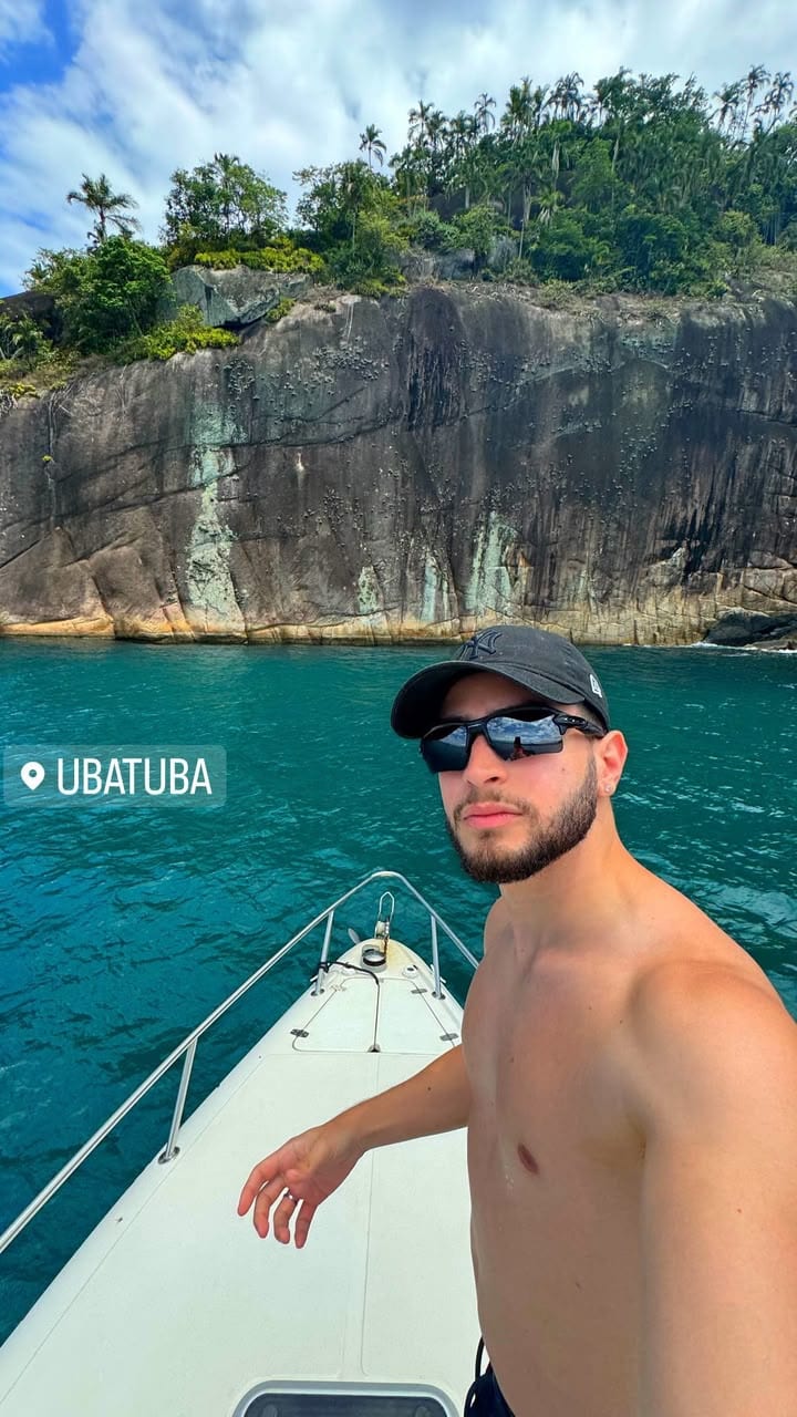 O Vagner sem camisa, de boné e óculos escuros, na proa de uma lancha no mar verde de Ubatuba, com o costão de pedra e a mata atrás.