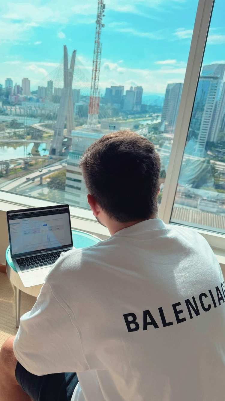 O Vagner de costas, trabalhando no notebook diante de uma janela alta, com a cidade e a ponte estaiada lá embaixo.