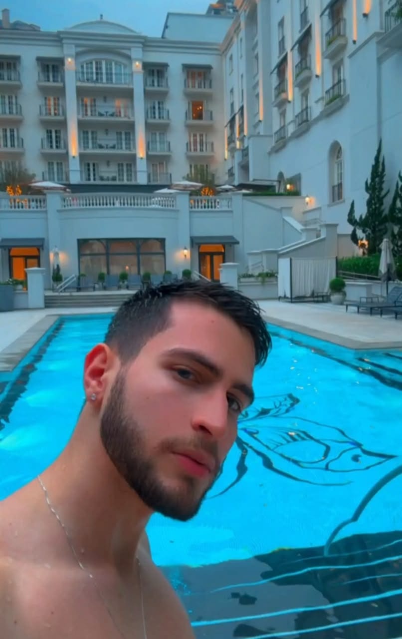 O Vagner de cabelo molhado, dentro da piscina de um hotel clássico, com a fachada iluminada ao anoitecer atrás.